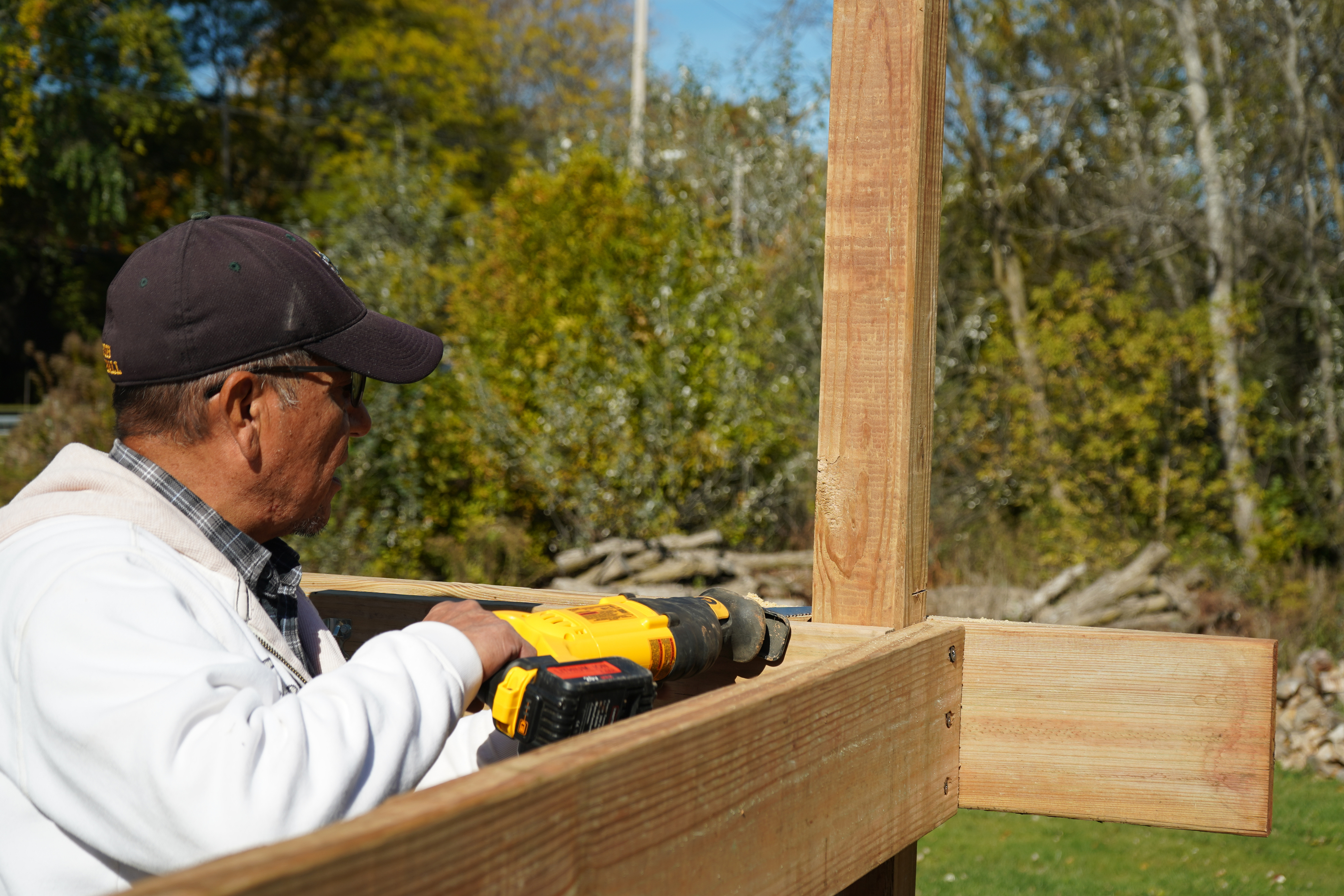 Person wearing a white sweatshirt using a handheld electric tool to construct a wooden frame. Bushes and trees are in the background.