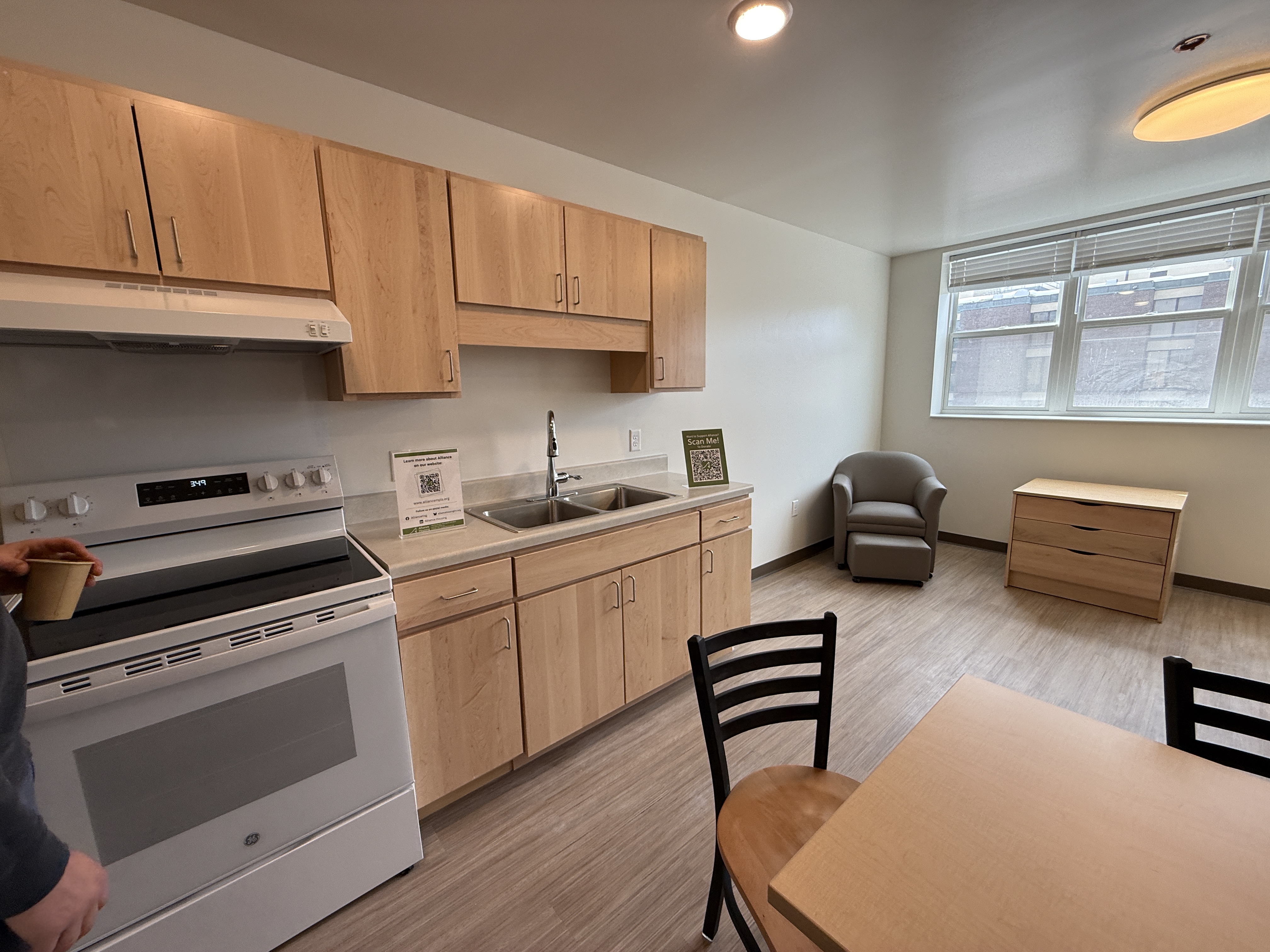 Modern apartment kitchen with light wood cabinets, a small dining room table, two chairs, and a window along the back wall.