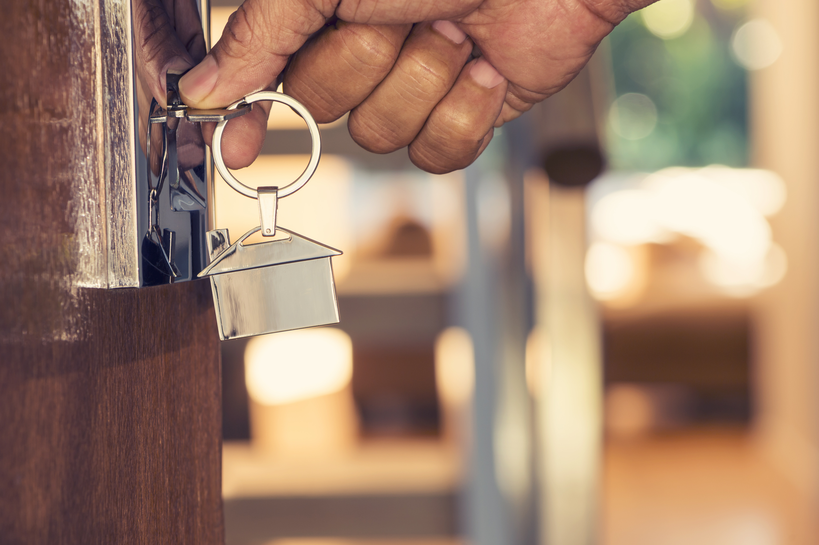 Hand opening a wooden door with a silver key and house-shaped keychain. The background is a blurred home interior.